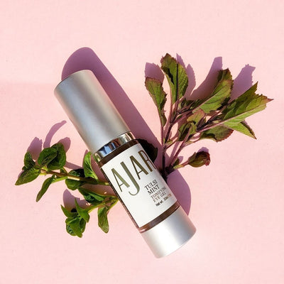 Tulsi Mint Tonifying Eye Gel bottle with a tulsi twig beneath it and a pink background
